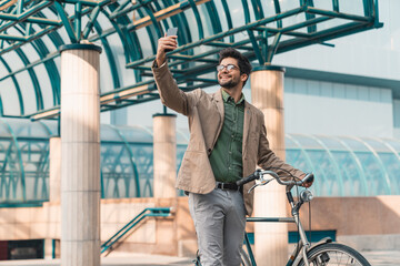 Cheerful happy handsome man with glasses taking selfie on the bike in front of business building.