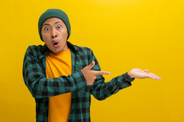 An excited young Asian man, wearing a beanie hat and casual shirt, points at his open palm...