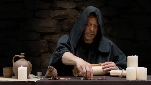 Monk counting money in stacks sitting at table in cell, dark tone.