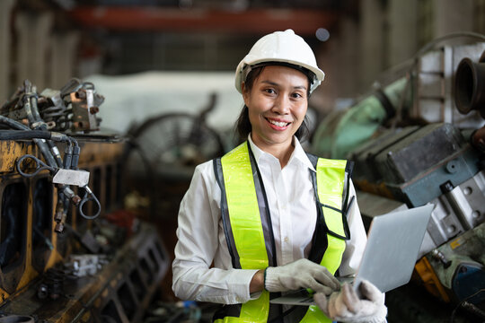 Confident Asian female engineer with labtop computer smiles while wearing a white hard hat and reflective vest in a mechanical workshop.