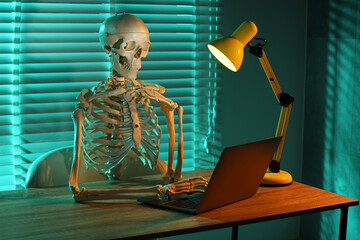 Waiting concept. Human skeleton sitting at wooden table with laptop indoors