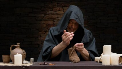 A monk pours money into a leather wallet while sitting at a table in a cell against a stone wall.