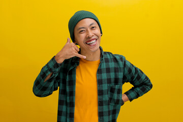 Attractive young Asian man, dressed in a beanie hat and casual shirt, is gesturing a CALL ME sign with his hand while standing against a yellow background