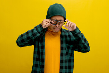 Asian man in a beanie and plaid shirt rubs his irritated eyes, pinching his nose in discomfort. He...