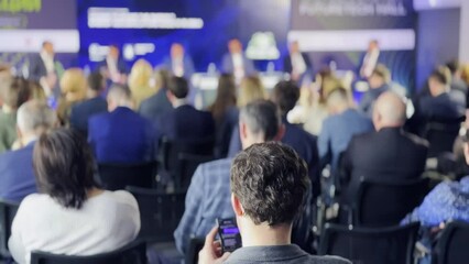 Rear view of diverse business professionals seated at a technology conference, listening attentively to speakers on stage.