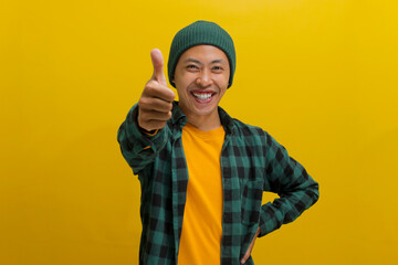 Confident Asian man is expressing approval and recommendation by showing a thumbs-up gesture, indicating his approval or liking for something positive. Standing against yellow background.
