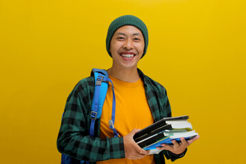 A confident young Asian student, dressed in a beanie hat and casual shirt and carrying a backpack, looks happy while holding a pile of books and standing against a yellow background