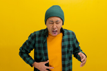 Displeased young Asian man, dressed in a beanie hat and casual shirt, is sticking out his tongue in...