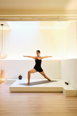 A woman is doing Virabhadrasana II or Warrior II yoga pose in a room with a white wall and a wooden floor. The room is dimly lit, and there is a lamp on the floor.