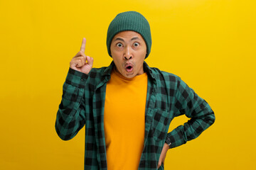 Excited young Asian man, dressed in a beanie hat and casual shirt, has an idea, raising his finger...