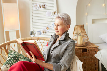 Side view of elegant stylish aged female with short haircut in gray denim jacket reading...