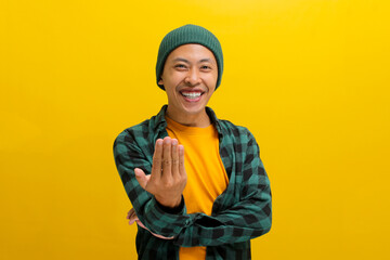 Happy, smiling young Asian man, dressed in a beanie hat and casual shirt, invites with a beckoning COME HERE gesture while standing against a yellow background.