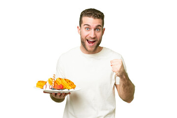 Young handsome blonde man holding waffles over isolated chroma key background celebrating a victory...