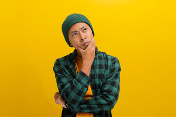 Fototapeta premium Thoughtful young Asian man, dressed in a beanie hat and casual shirt, appears concerned as he rests his hand on his chin, contemplating about the future while standing against yellow background.