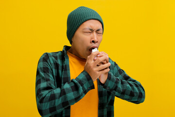 Young Asian man is sneezing and blowing his nose into a napkin, showing signs of suffering from a...