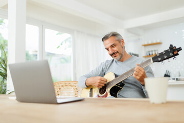Middle-aged man enjoys playing guitar while working from home, laptop open on a bright, modern...