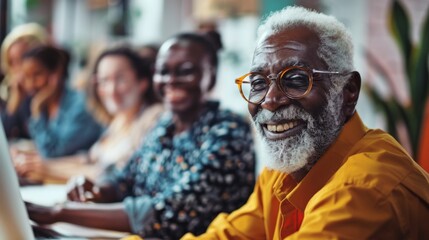 Cheerful elderly man with white hair and a full beard wearing glasses and a yellow shirt sitting at a table with a laptop surrounded by blurred figures of people