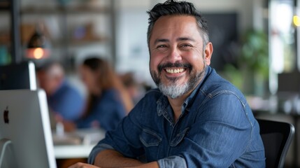 Obraz premium Smiling man with gray beard and hair wearing blue denim shirt sitting at desk with laptop in modern office environment with blurred background of colleagues and plants.