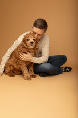 Obraz premium Studio portrait of a handsome caucasian man in his 40s sitting on the floor and embracing his cocker spaniel dog. The background is beige. The man is wearing jeans and beige shirt. 