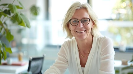 Smiling woman with blonde hair and glasses wearing a white blouse and gold necklace sitting at a desk with plants in the background.
