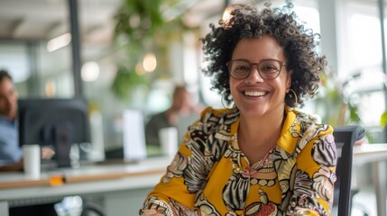Smiling woman with curly hair wearing glasses sitting in an office with a yellow and white patterned blouse.