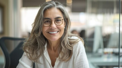 Smiling woman with glasses wearing white blouse sitting in office with blurred background.