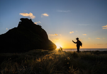 Man taking photos using a smartphone of Lion Rock at sunset. Unrecognizable kids playing on Piha...