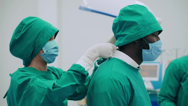 Team of professional multiracial surgeons getting dressed preparing to operation. Nurses helping chief surgeon to put on the protective robe before surgery in operating room at hospital.