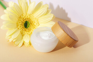 Cosmetic cream jar and yellow gerbera flowers on neutral background in sunlight. Closeup