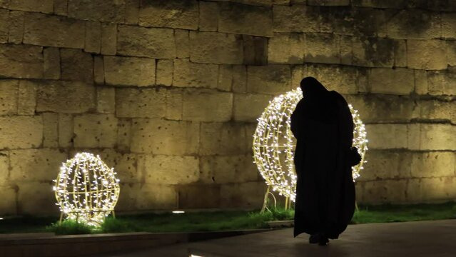Traditional women's outerwear in Muslim countries. A woman in a black niqab walks at night along a lighted street next to an ancient stone wall. A religious woman. Muslim robes. Lamps outside at night