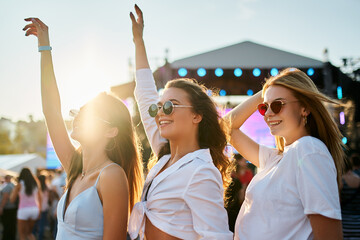 Group of joyful young women dance at sunny beach music festival. Girls in summer outfits enjoy concert, hands up in celebration. Vibrant fest atmosphere, friends share fun experience by sea.