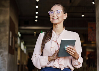 An optimistic businesswoman in glasses looks upward while holding a tablet, standing in a modern...
