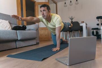 Fototapeta premium Man in quadruped position stretches arm and opposite leg with effort on yoga mat, accompanied by laptop and sleeping black cat in the couch.
