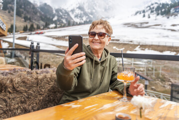 Attractive senior woman traveler relaxing in a cafe at a ski mountain resort drinking a cocktail and using smartphone