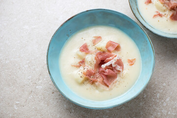 Potato soup served with torn prosciutto in a turquoise bowl, horizontal shot on a beige stone background, selective focus