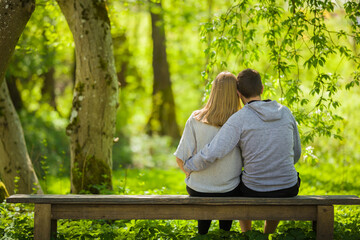 Young couple hugging each other, sitting on bench and staring at green leaves, grass and trees at beautiful park in sunny spring day. Spending time in nature. Peaceful atmosphere. Back view.