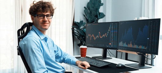 Profile of smiling young stock trader's curly hair looking at camera, sitting against on financial exchange screen at office on panorama image. Concept of dynamic business digital investment. Gusher.