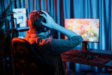Host channel of stressful young gaming streamer getting to lose in fighting Moba at battle arena defeated multiplay team, wearing headphone on pc monitor with back side image at red neon room. Gusher.