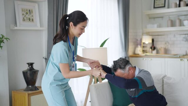 Young woman Asian nurse caretaker assists a senior man as he settles into a seat in the living room of a nursing home, ensuring his comfort and safety.