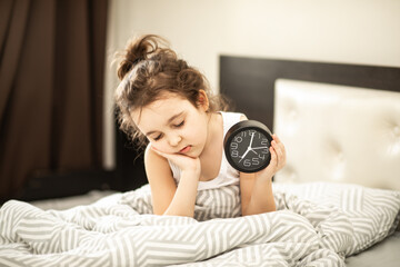 Cute little kid sleepy girl sitting on bed waking up in morning and holding alarm clock.