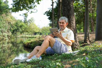 happy asian senior man relaxing under a tree in the park,using digital tablet communicate with family,concept of elderly retired with modern lifestyle,technology,travel,relax
