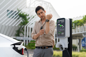Young man checking time on smartwatch while EV charger to recharging battery from charging station...