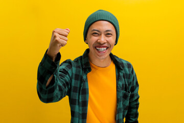 Excited young Asian man, dressed in a beanie hat and casual shirt, is expressing joy and celebrating victory with a fist pump or a YES gesture while standing against a yellow background