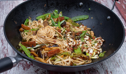 A delicious vegan stir fry consisting of seitan, eggplant, snow peas and other greens stir fried in steel wok, served along with noodles