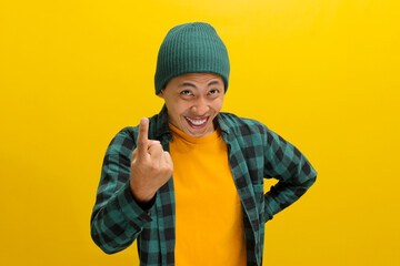 Happy, smiling young Asian man, dressed in a beanie hat and casual shirt, invites with a beckoning COME HERE gesture while standing against a yellow background.