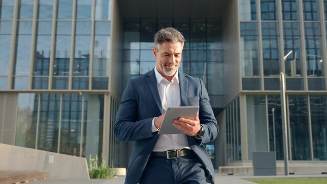 Hispanic older smiling man walking outdoor at business building using touchpad computer for work. Confident focused successful mature Indian or Latin entrepreneur businessman holding digital pc tablet