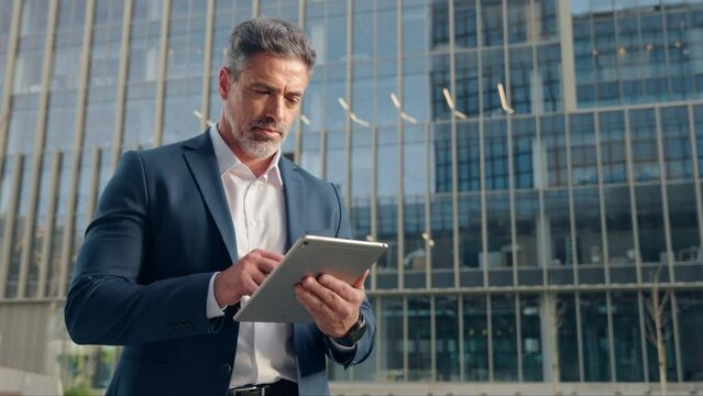 Confident focused successful mature Indian or Latin entrepreneur businessman holding digital pc tablet standing outdoor at business building. Hispanic older man in suit using touchpad computer at work