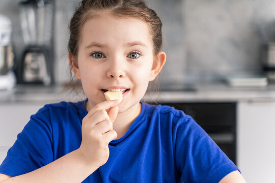 A beautiful little girl puts a piece of mozzarella cheese in her mouth. A pre-teen girl in a blue blouse against the background of the kitchen at home. Selected focus