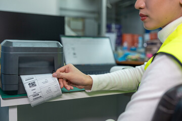 Beautiful Asian woman uses a laptop or notebook to print bar code stickers on a bar code printer. Selective focus at sticker barcode.