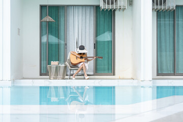 A young Asian woman, tourist, sits contentedly by the poolside of a luxury hotel room, strumming a guitar and singing, dressed in a stylish ensemble of dress, hat, and sunglasses, enjoying her summer